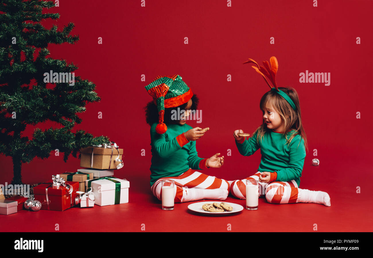 Petites filles, assis près d'un arbre de Noël avec des biscuits et du lait. Deux enfants heureux vêtus de vêtements thème Noël assis sur le plancher à côté d'un ch Banque D'Images
