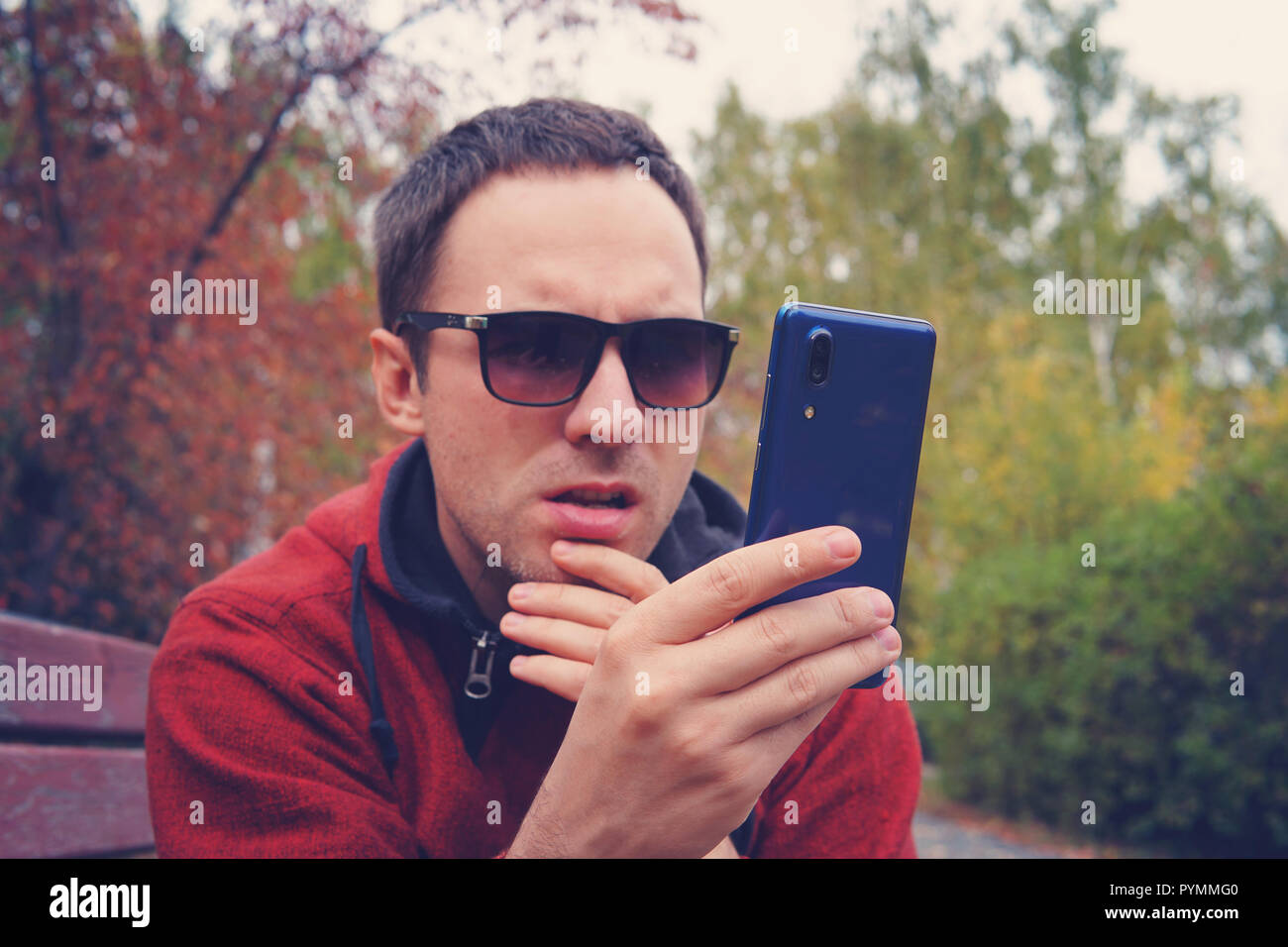 Jeune homme dans le port de lunettes est titulaire d'un smartphone moderne bleue dans sa main et regarder à l'écran avec un regard surpris. La perplexité sur le visage. L Banque D'Images