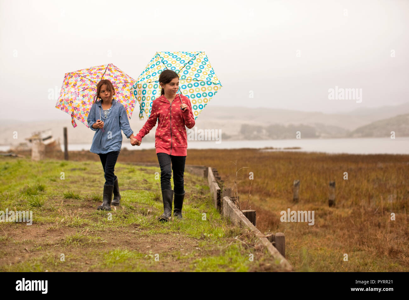 Deux jeunes filles nous tenir la main comme ils portent des parapluies ouverts et marcher le long d'un chemin herbeux à proximité d'une terre marécageuse. Banque D'Images