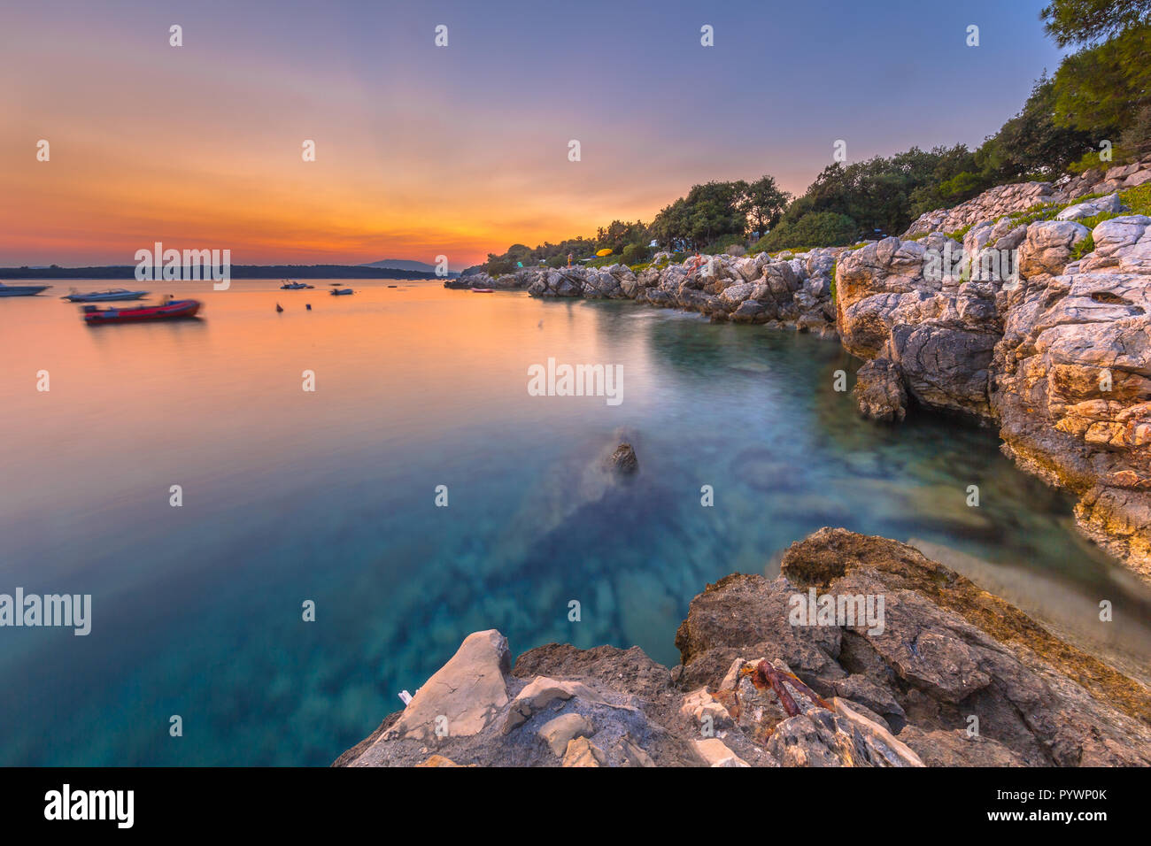 De soleil colorés sur la côte rocheuse de la Croatie. Une longue exposition de droit de coucher du soleil, les rochers, les bateaux et l'eau turquoise de l'île de Cres. Banque D'Images De soleil colorés sur la côte rocheuse de la Croatie. Une longue exposition de droit de coucher du soleil, les rochers, les bateaux et l'eau turquoise de l'île de Cres. Banque D'Images