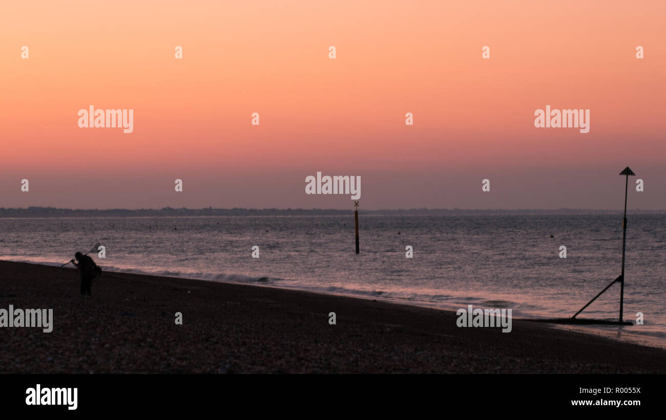 Homme avec détecteur de métal enthusiast transporter son détecteur de métal sur une plage de galets au lever du soleil Banque D'Images