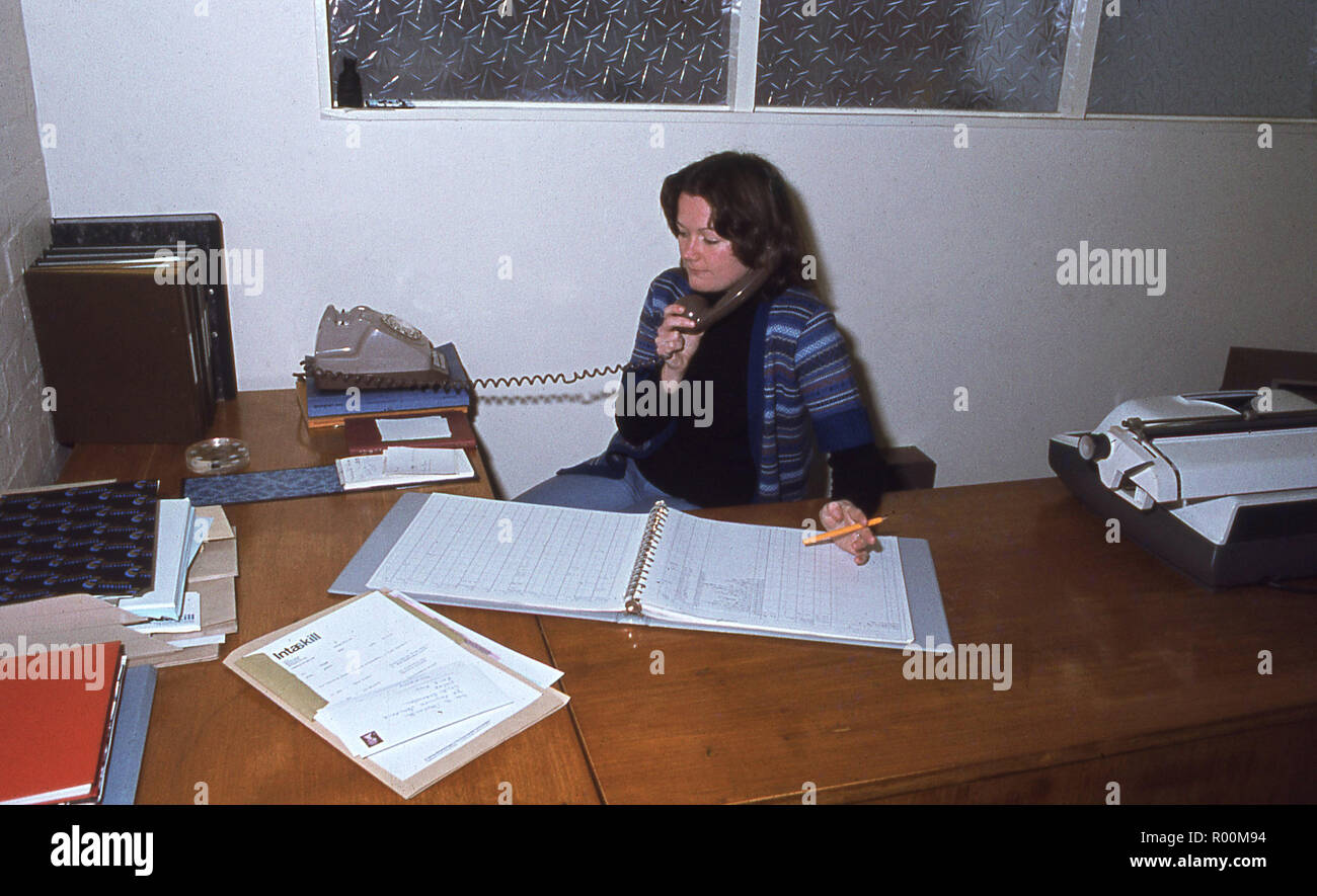 Années 1970, female office worker secrétaire ou à son bureau parlant au téléphone, avec les dossiers de l'entreprise ou les figures dans un dossier ouvert devant elle, England, UK. Banque D'Images