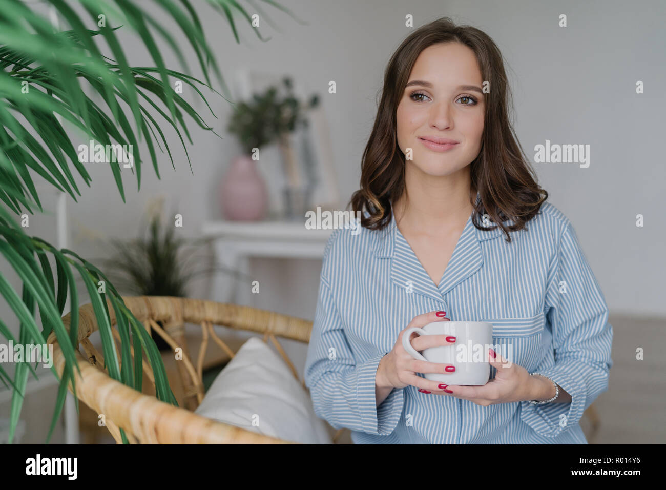 Tourné à l'intérieur du plaisir jolie femme avec les cheveux foncés, une peau saine, boit du café le matin, porte pyjama, l'air songeur de côté, pose à Banque D'Images