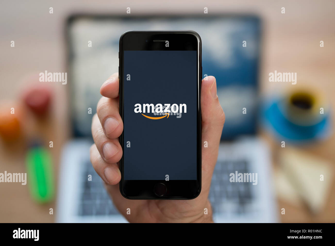 Un homme se penche sur son iPhone qui affiche le logo d'Amazon, tout en s'assit à son bureau informatique (usage éditorial uniquement). Banque D'Images