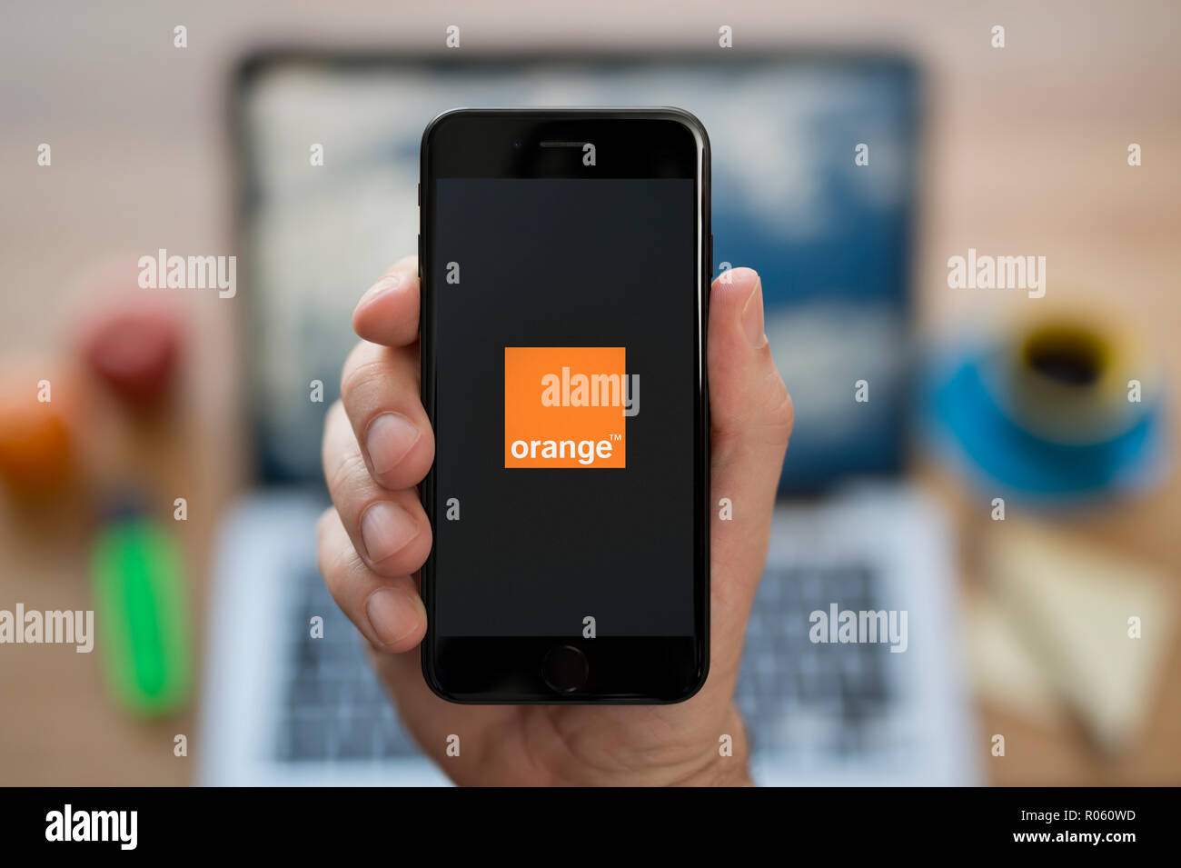Un homme se penche sur son iPhone qui affiche le logo Orange, tandis qu'assis à son bureau de l'ordinateur (usage éditorial uniquement). Banque D'Images