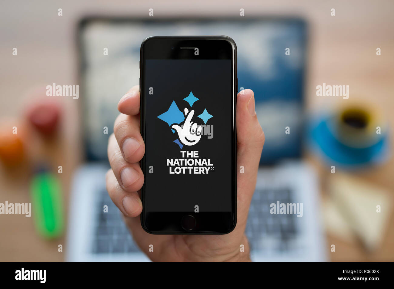 Un homme se penche sur son iPhone qui affiche le logo de la loterie nationale, tout en s'assit à son bureau informatique (usage éditorial uniquement). Banque D'Images