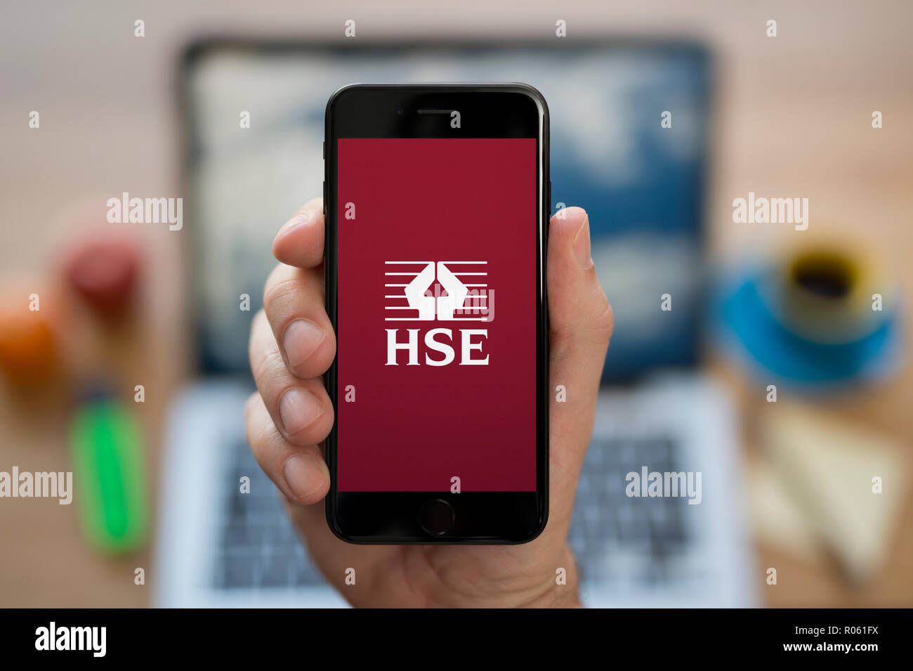 Un homme se penche sur son iPhone qui affiche le Health and Safety Executive (HSE), tandis que le logo était assis à son bureau de l'ordinateur (usage éditorial uniquement). Banque D'Images