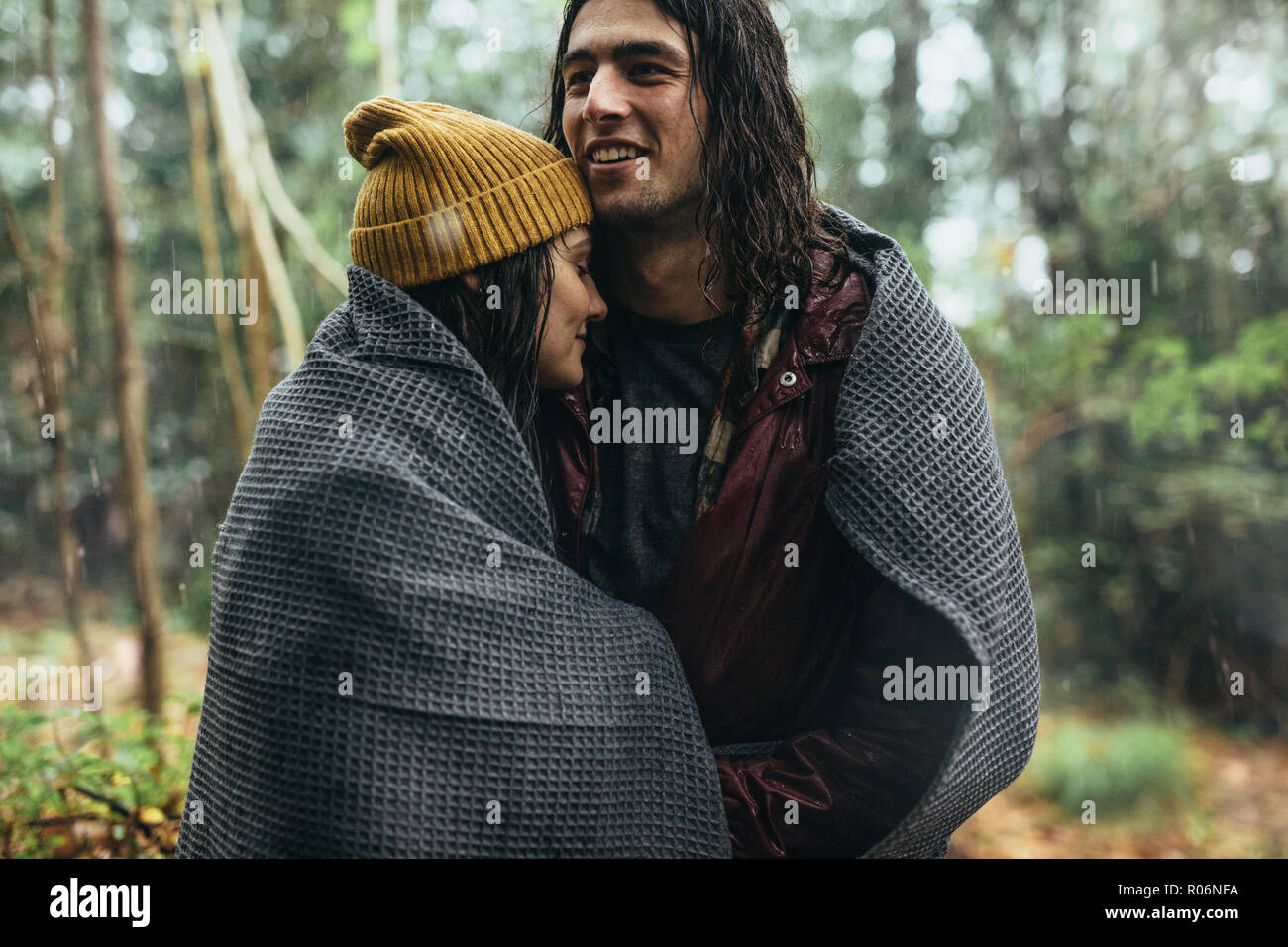 Couple dans le contrat cadre sous la pluie à l'extérieur. Jeune homme et femme enveloppée dans un plaid debout sous la pluie au parc. Banque D'Images