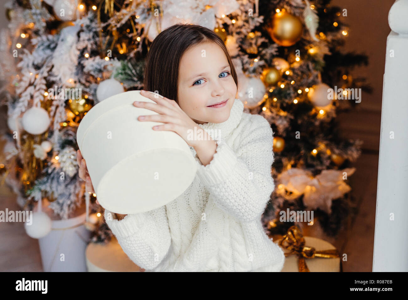 Blue Eyed joli joli petit enfant tient présent fort, se demande ce qu'est à l'intérieur, se dresse près de l'arbre de Noël ou Nouvel An, reçoit des parents. surprise Banque D'Images