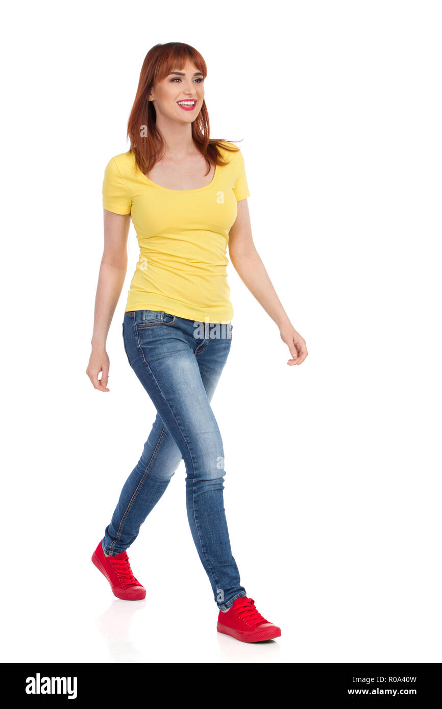 Jeune femme en chemise jaune, jeans et baskets rouge est la marche, souriant et à l'hôtel. Vue latérale avant. Studio pleine longueur shot isolé sur blanc. Banque D'Images
