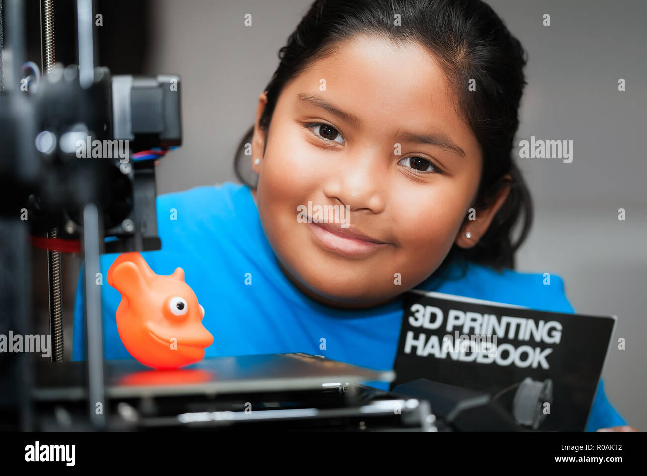 Cute girl smiling et montrant sa création imprimée en 3d sur le lit d'une imprimante 3D tout en maintenant une brochure qui enseigne à propos de l'impression 3d. Banque D'Images