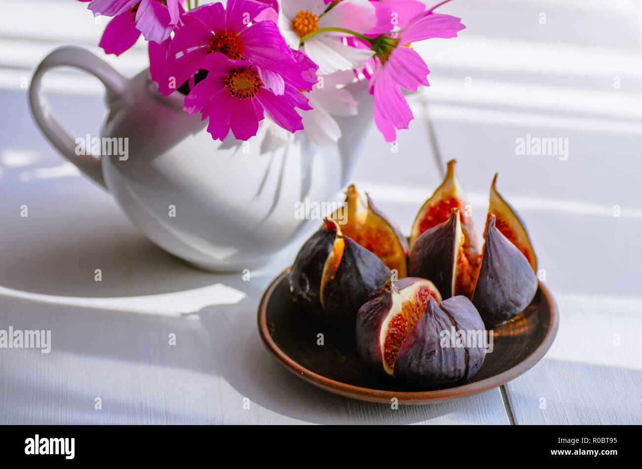 Vase blanc avec des fleurs. Figues mûres dans une assiette sur un fond de bois blanc. Journée ensoleillée. Banque D'Images