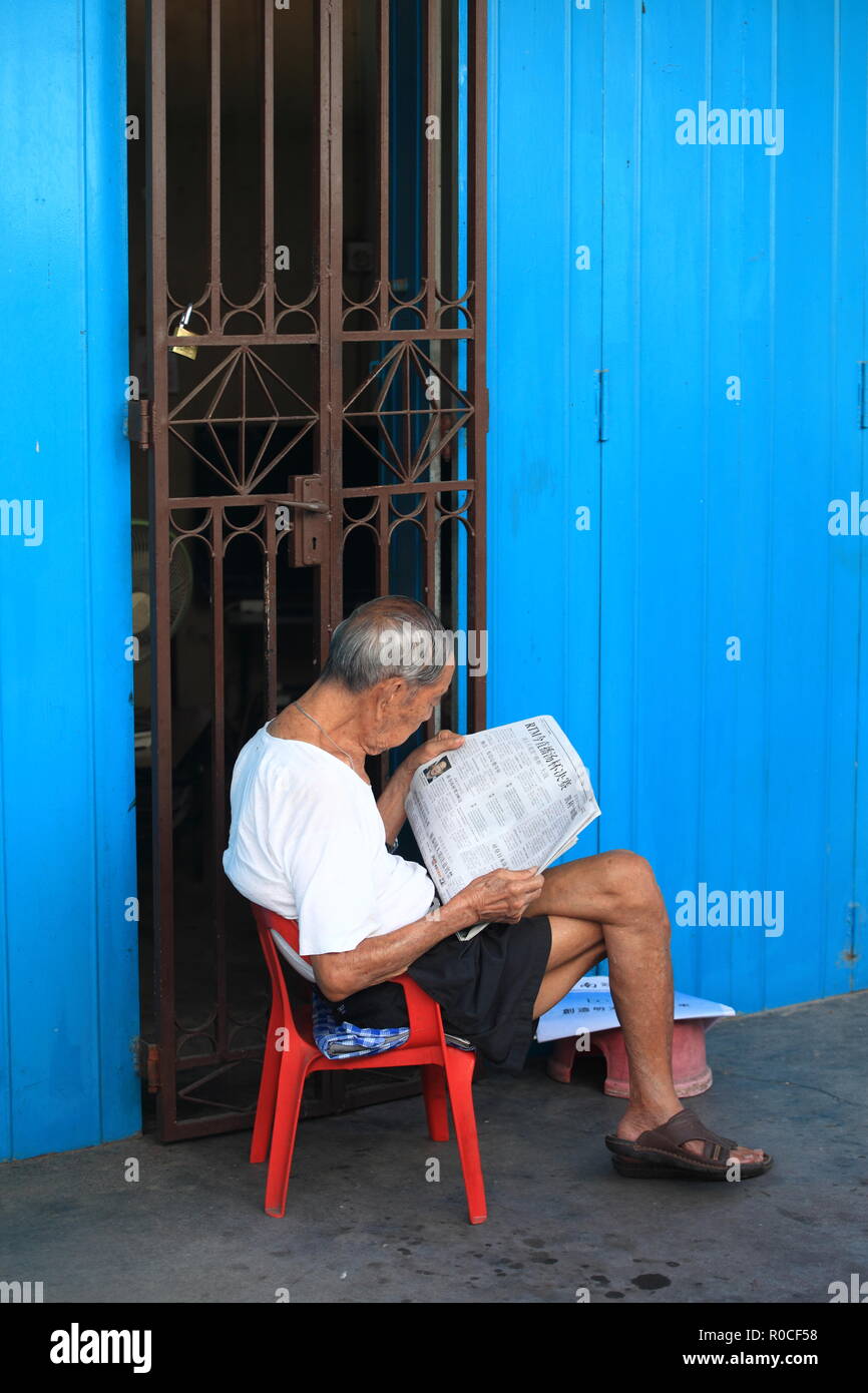 Un vieil homme chinois local en chemise blanche basique et sandales, lisant le journal local sur une courte chaise en plastique rouge Melaka, Malaisie Banque D'Images