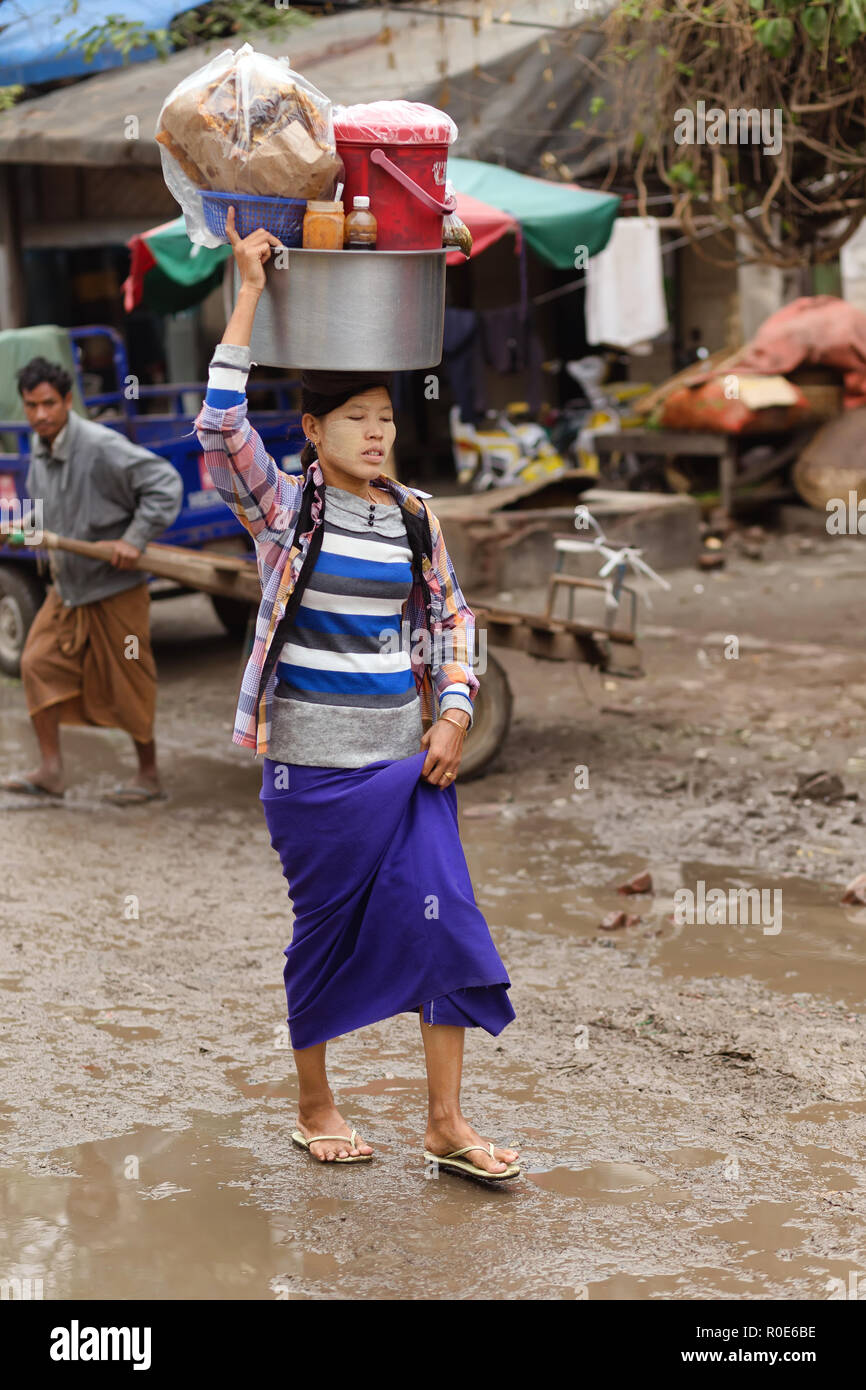 AMARAPURA, Myanmar, le 17 janvier 2015 : une femme est porteur d'un grand conteneur métallique sur la tête, marchant dans la rue sale du marché Zegyo, dans Banque D'Images