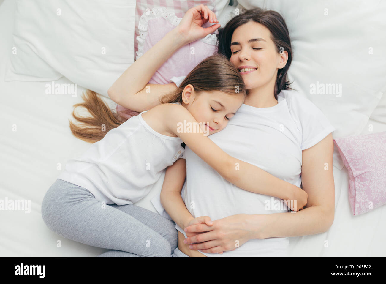 Vue de dessus de cheerful mère dort sur un lit blanc près de sa fille qui  embrasse maman avec beaucoup d'amour, montre une bonne attitude,  d'agréables rêves et de nice Photo Stock -