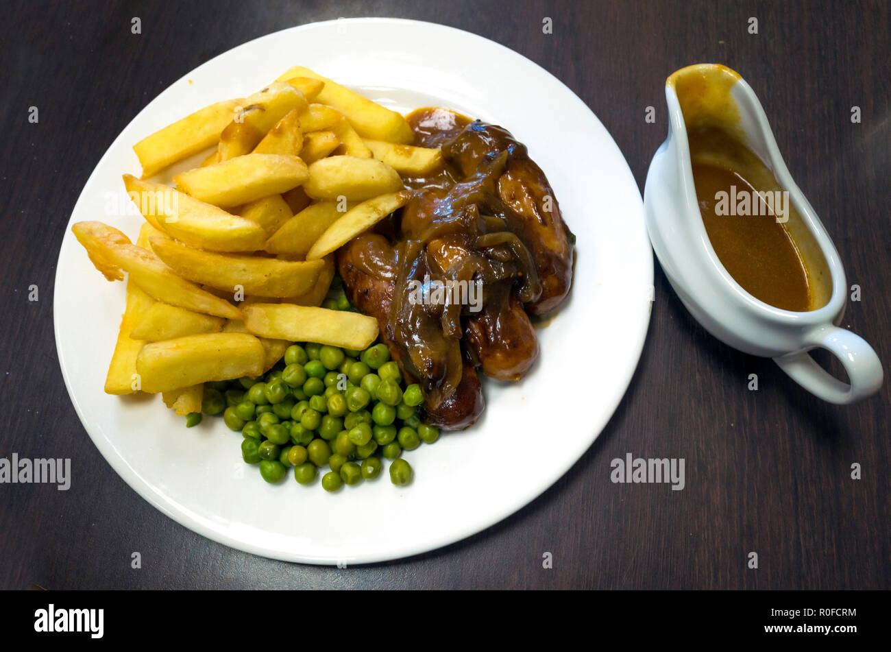 De savoureux repas de midi et les oignons saucisses Yorkshire pucer pommes de terre, pois et un petit pot de sauce moutarde Banque D'Images