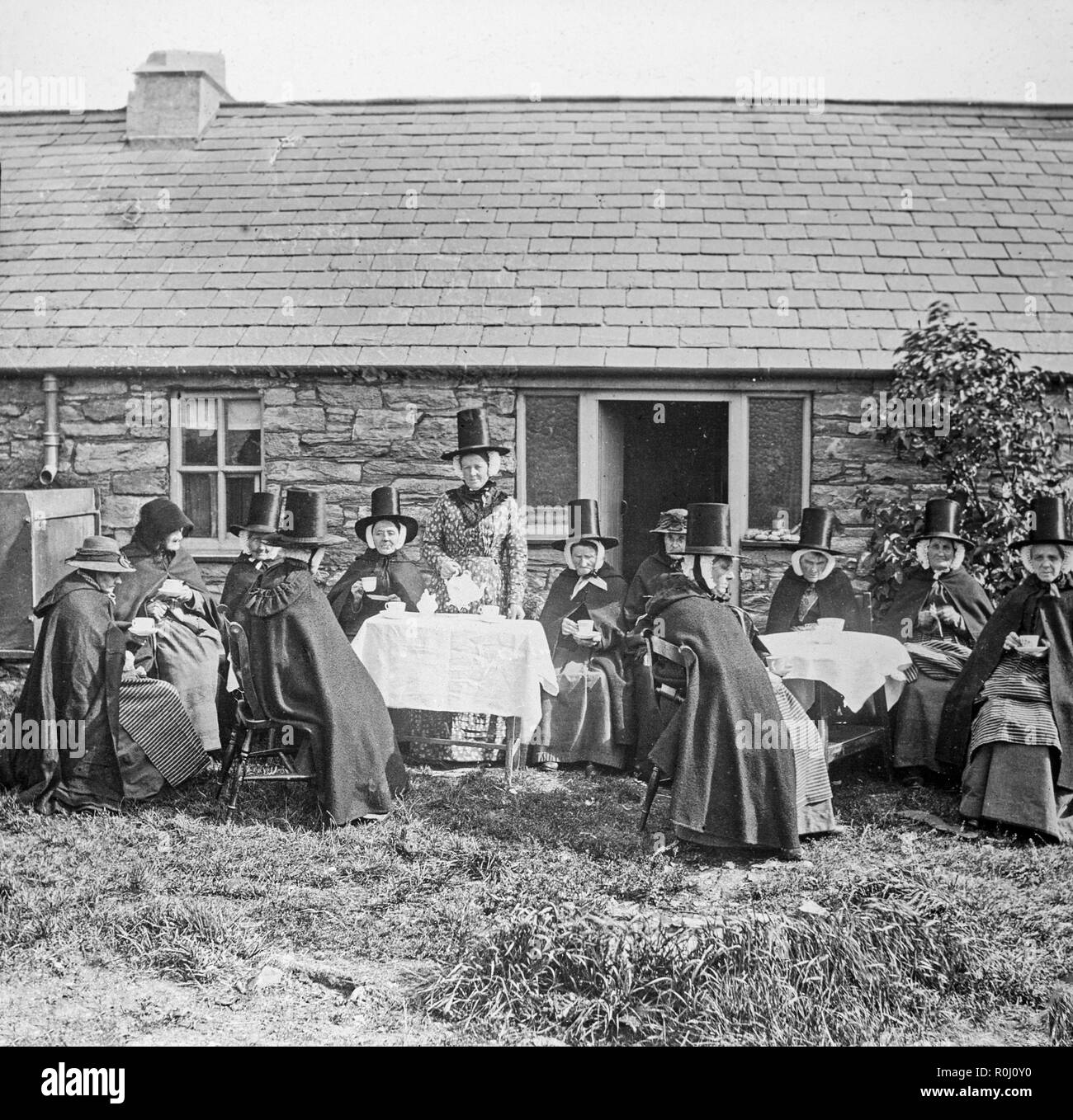 Alate Victorian vintage photographie noir et blanc, montrant un groupe de femmes habillées en costume traditionnel gallois, en dehors d'une petite maison. Certains boivent le thé, certains font de la couture. Le chapeau traditionnel Gallois est très bien illustré. Banque D'Images