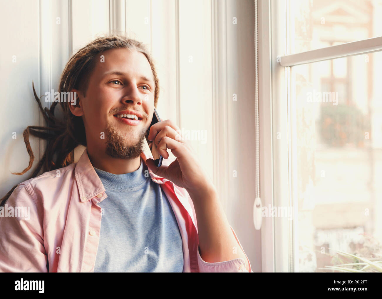 Homme barbu attrayant Hipster avec dreads portant des écouteurs assis près de la fenêtre et using smartphone Banque D'Images