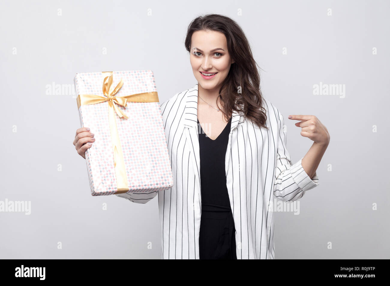 Portrait de jeune femme brune magnifique satisfait en blouse blanche l'article montre de l'index et de présenter avec bow jaune et sourire à pleines dents,recherche à Banque D'Images Portrait de jeune femme brune magnifique satisfait en blouse blanche l'article montre de l'index et de présenter avec bow jaune et sourire à pleines dents,recherche à Banque D'Images