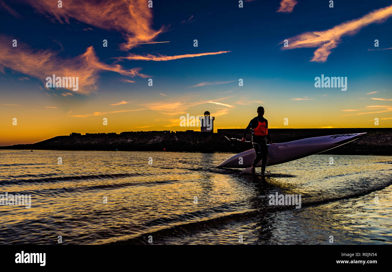 Un homme qui sort de la mer portant un kayak au coucher du soleil, un phare et le ciel bleu en arrière-plan. Carmarthenshire, Pays de Galles. UK. Banque D'Images