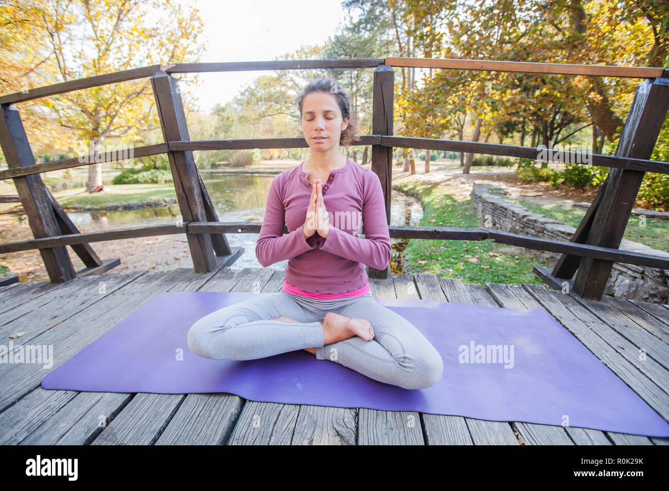 Belle jeune femme dans une pose méditative sur pont en bois au parc. Femelle en bonne santé faisant de l'exercice de yoga, Lotus padmasana pose, matin ensoleillé d'automne. Banque D'Images