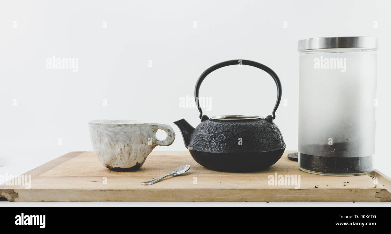 Théière en fonte avec cannister de thé noir et de céramique faite à la main / tasse mug sur un plateau en bois pâle fond blanc avec des prix pour la copie. Hygge. Banque D'Images