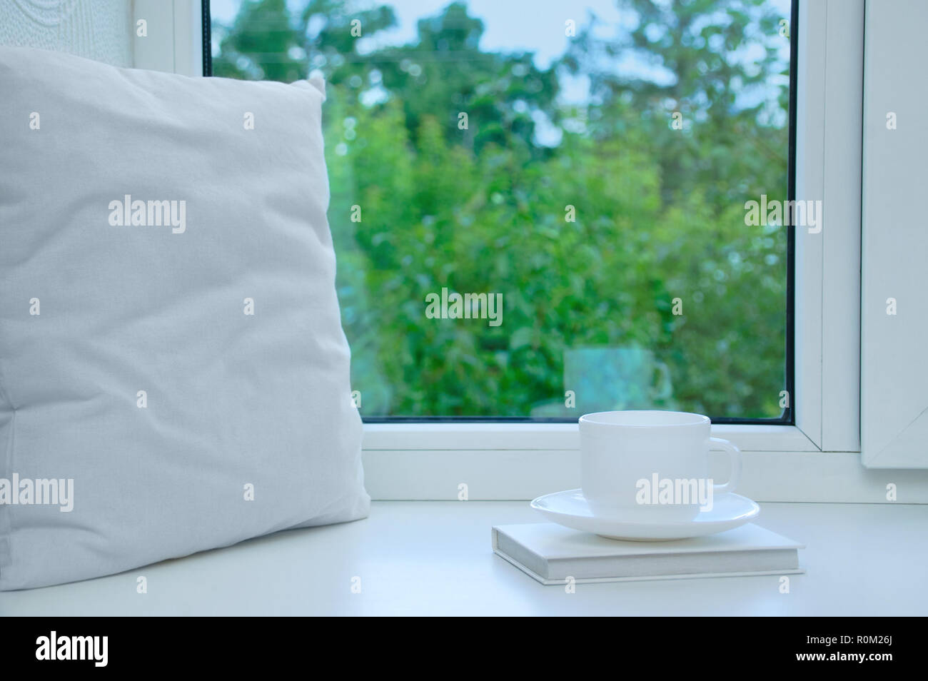 Un oreiller blanc, un livre et une tasse et soucoupe debout sur le rebord de la fenêtre dans l'été. Banque D'Images