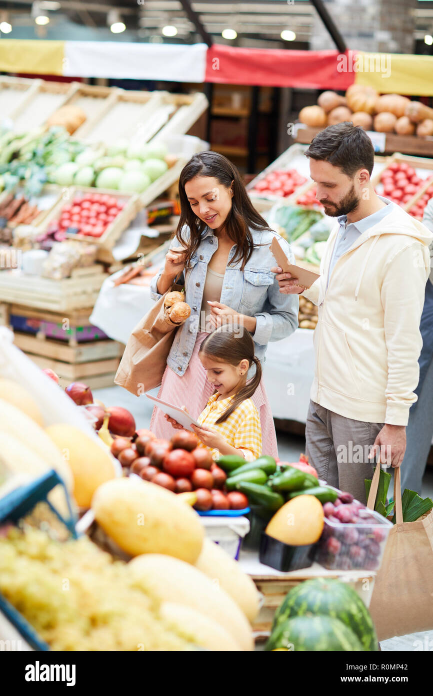 Jeune famille positive de l'alimentation à l'aide d'étagères et de magasinage en ligne pour l'achat d'application sur tablette, jolie fille à l'aide d'appareil moderne dans les aliments biologiques s Banque D'Images