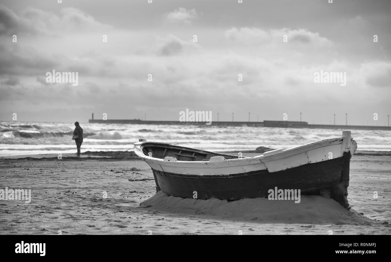 Moody photo noire et blanche d'un bateau abandonné coincé dans le sable d'une plage avec une seule personne dans la distance près de la rive avec des vagues et des nuages Banque D'Images
