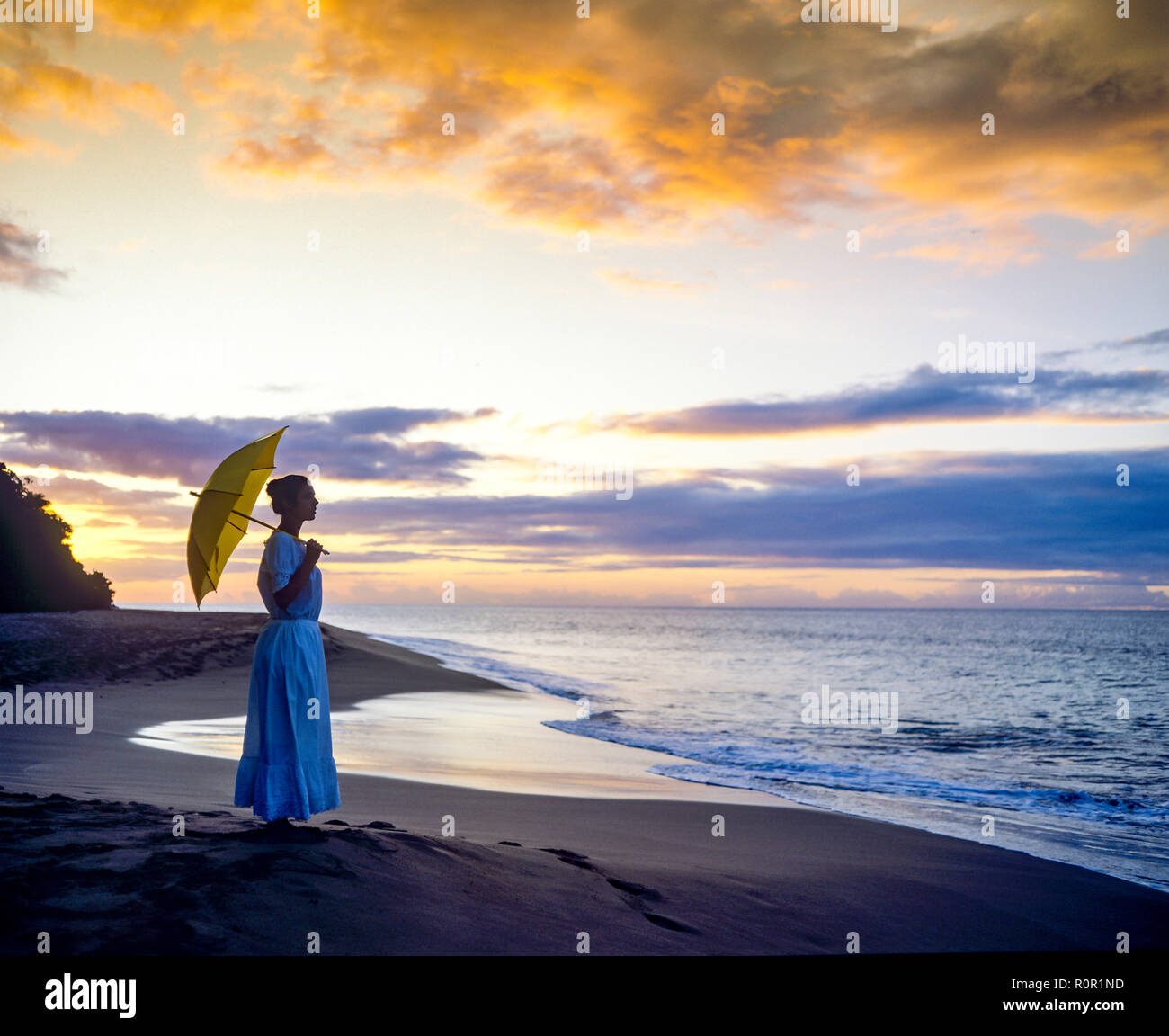 Jeune femme d'ossature avec robe longue et ombrelle face à la mer des Caraïbes au coucher du soleil, de la plage tropicale, Guadeloupe, French West Indies, Banque D'Images