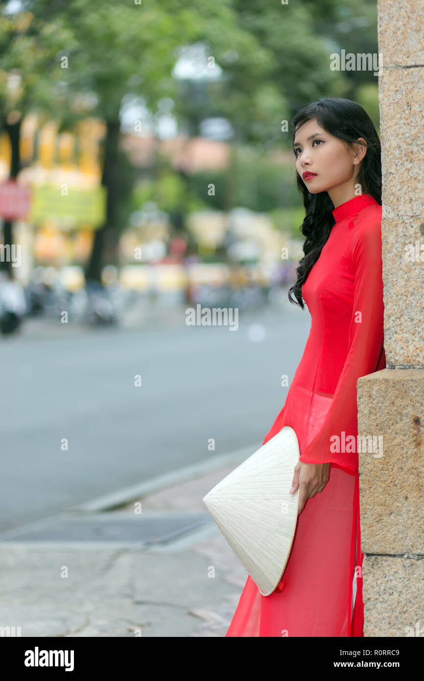 Superbe fille en vêtements traditionnels vietnamiens article appuyé contre un mur de briques dans une rue urbaine rêveusement regarder en l'air, side view Banque D'Images