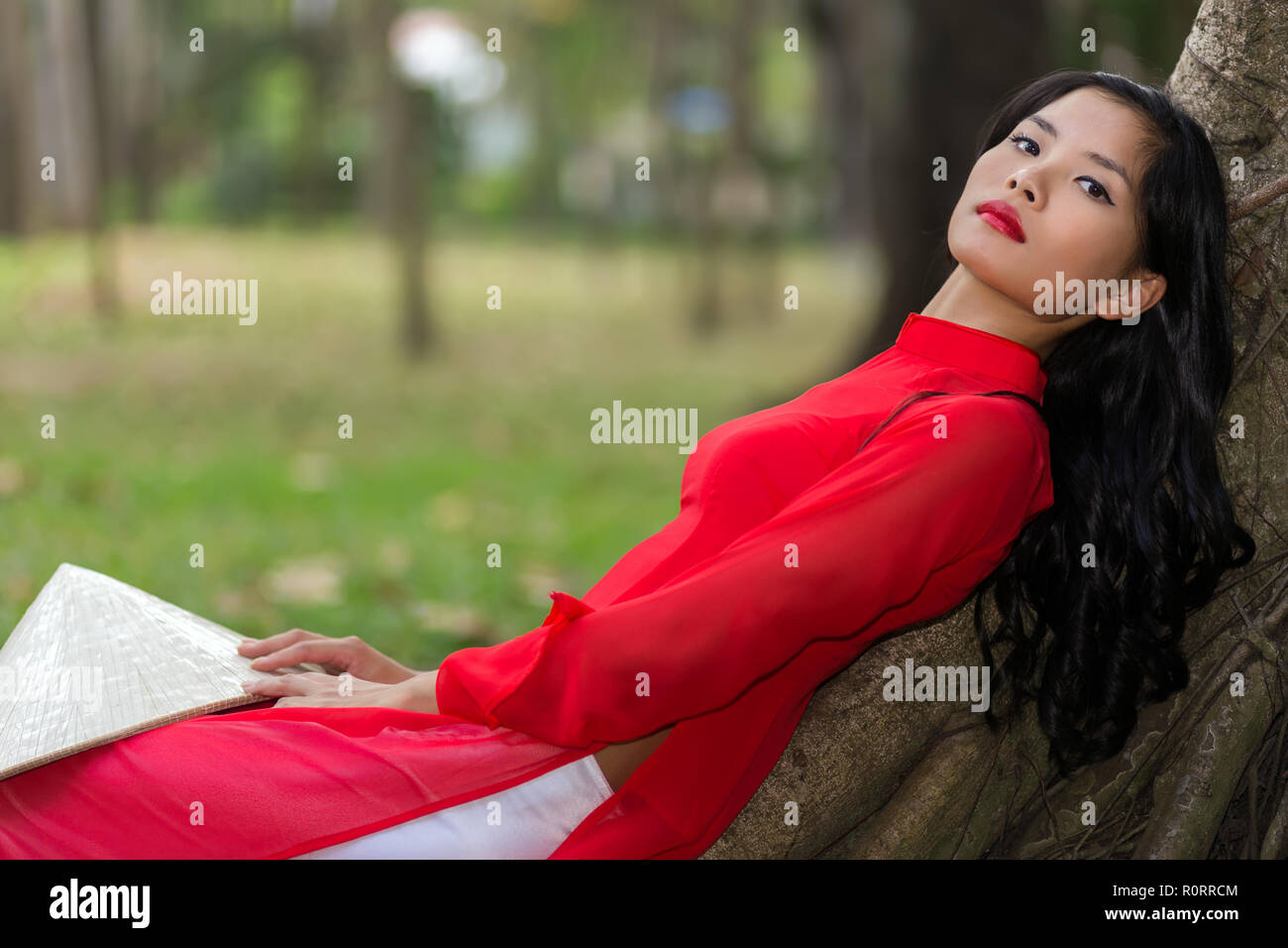 Jeune femme mince vietnamien de détente dans un parc en se penchant en arrière contre les racines aériennes d'un arbre dans son costume rouge traditionnel à la recherche de l'appareil photo Banque D'Images