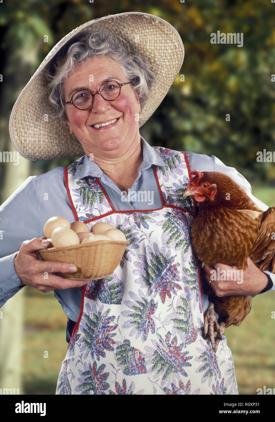 La femme d'un agriculteur portant des lunettes et un chapeau de paille  tient une poule rouge et un panier d'œufs fraîchement pondus. Elle a des  joues roses, des cheveux gris et un
