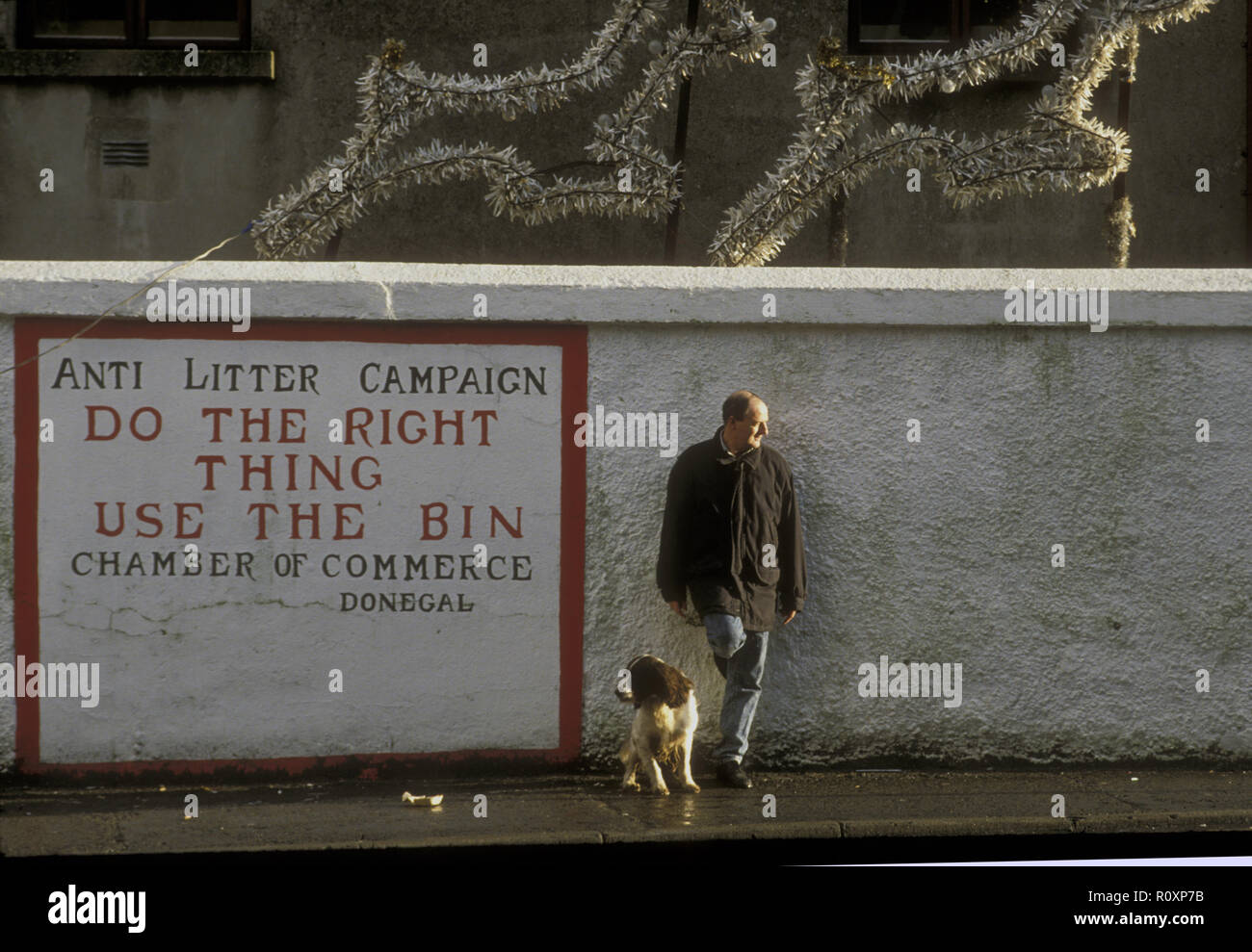 Un homme se tient immobile et se penche sur un mur à côté d'un panneau de campagne anti-litière. Son chien spaniel est assis à ses côtés. Au-dessus d'eux sont des décorations de Noël. Donegal, Irlande Banque D'Images