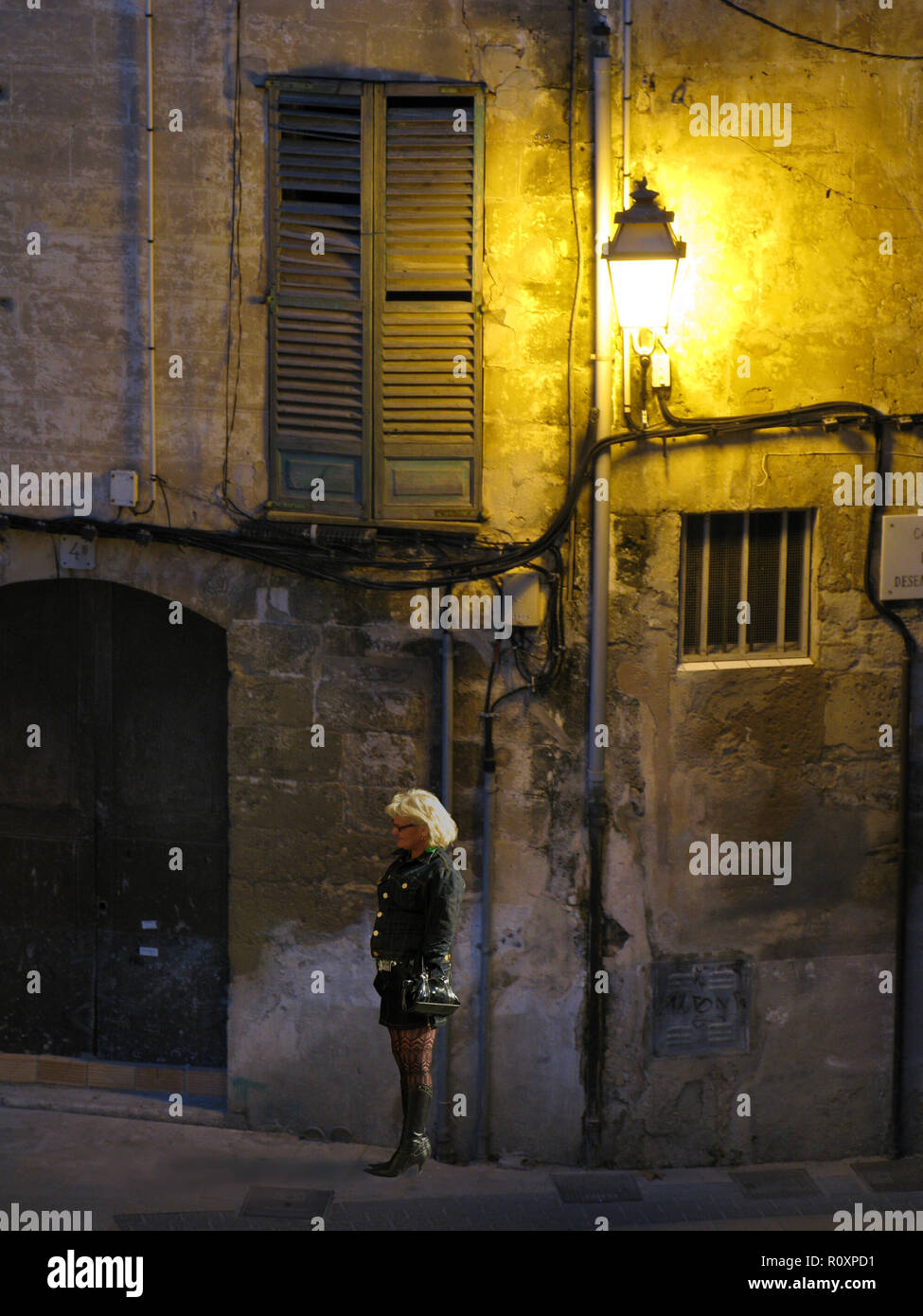 Une femme blonde en bottes, bas et jupe courte attend dans un coin de rue  sous une lumière de rue et de vieux volets dans le quartier Avenidas de  Palma, Majorque, Espagne