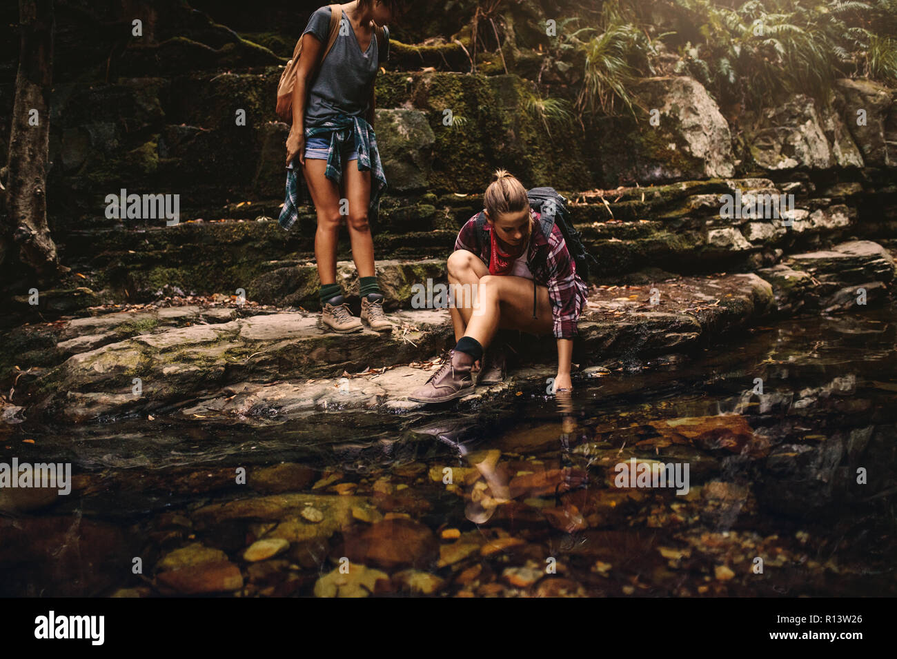 Deux femmes les randonneurs par un étang d'eau en montagne. Femme assise sur rocher et de mettre sa main dans l'eau avec l'ami par et à la recherche. Banque D'Images