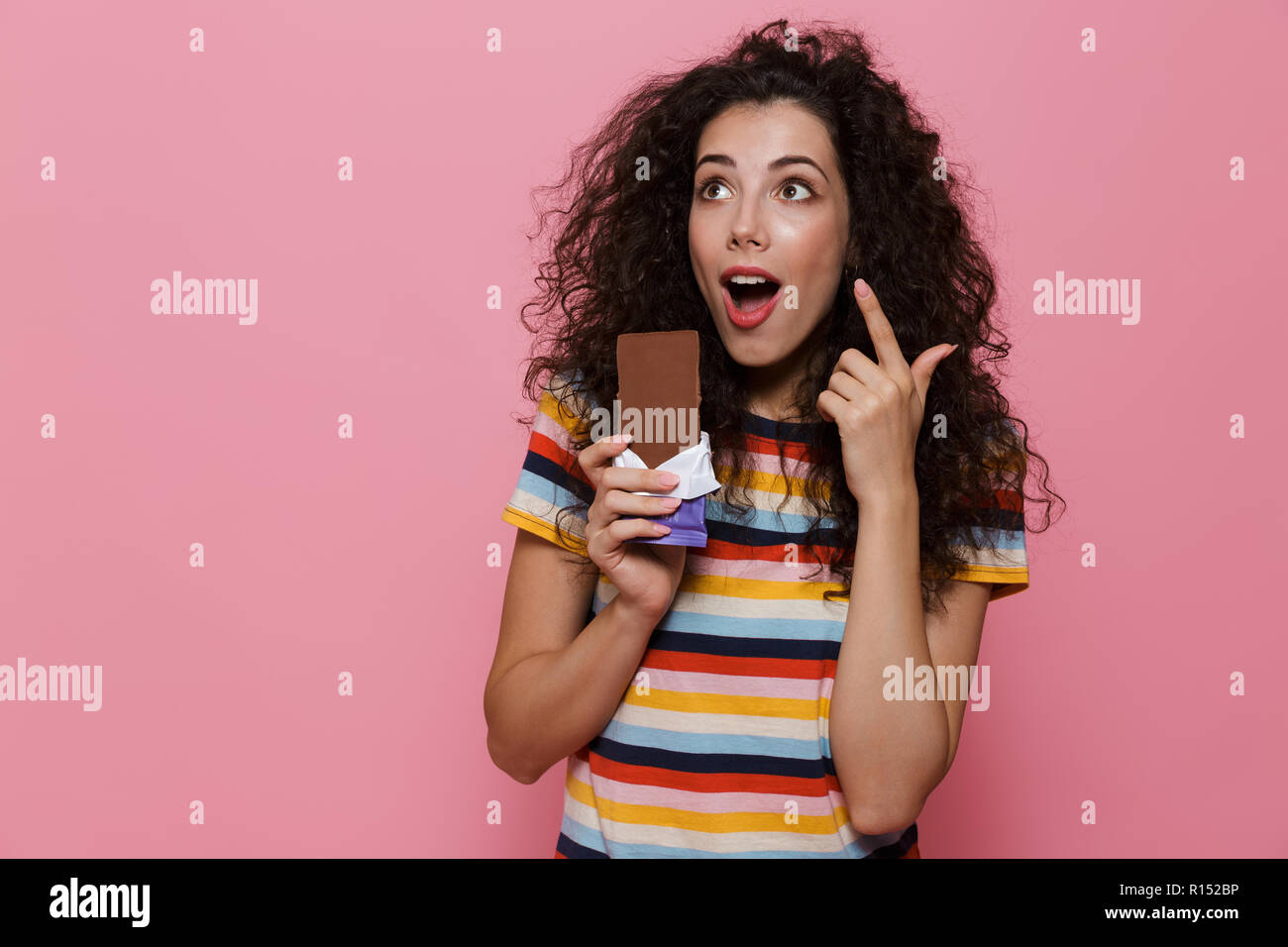Photo de jolie femme 20s avec des cheveux bouclés tenant une barre de chocolat plus isolé sur fond rose Banque D'Images
