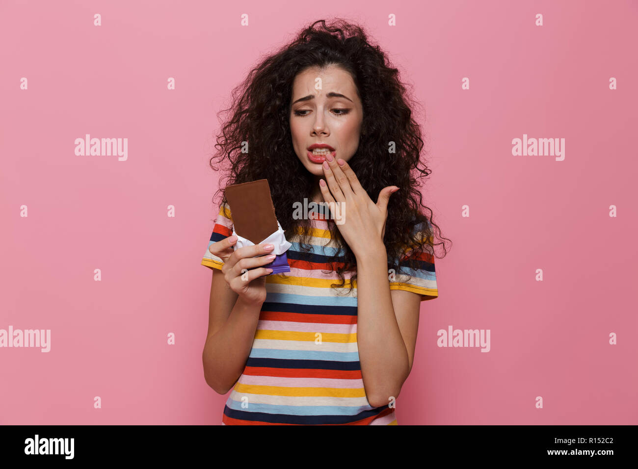Photo de femme confus 20s avec des cheveux bouclés tenant une barre de chocolat plus isolé sur fond rose Banque D'Images