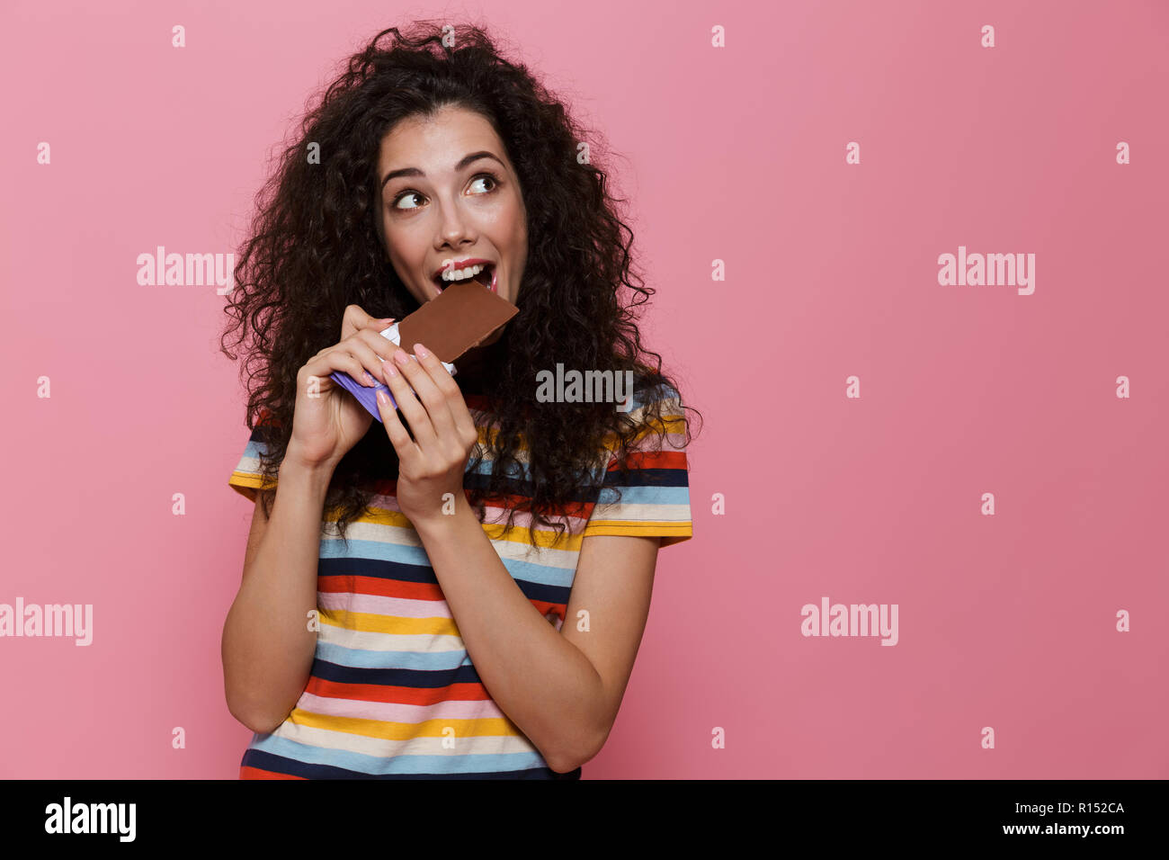 Photo de jeune femme de la vingtaine avec des cheveux bouclés manger du chocolat plus isolé sur fond rose Banque D'Images