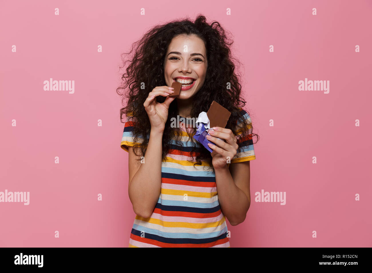 Photo de belle femme 20s avec les cheveux bouclés de manger une barre de chocolat plus isolé sur fond rose Banque D'Images