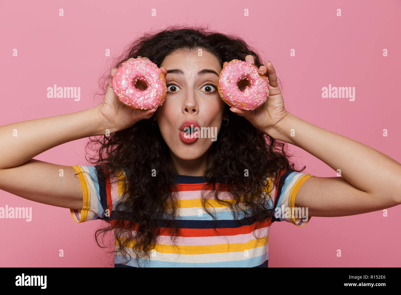 Photo de belle femme 20s avec les cheveux bouclés s'amusant et holding donuts plus isolé sur fond rose Banque D'Images