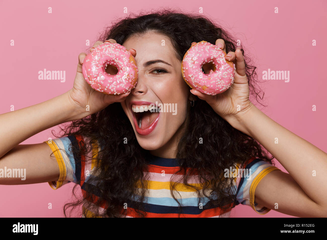 Photo de jolie femme 20s avec les cheveux bouclés s'amusant et holding donuts plus isolé sur fond rose Banque D'Images