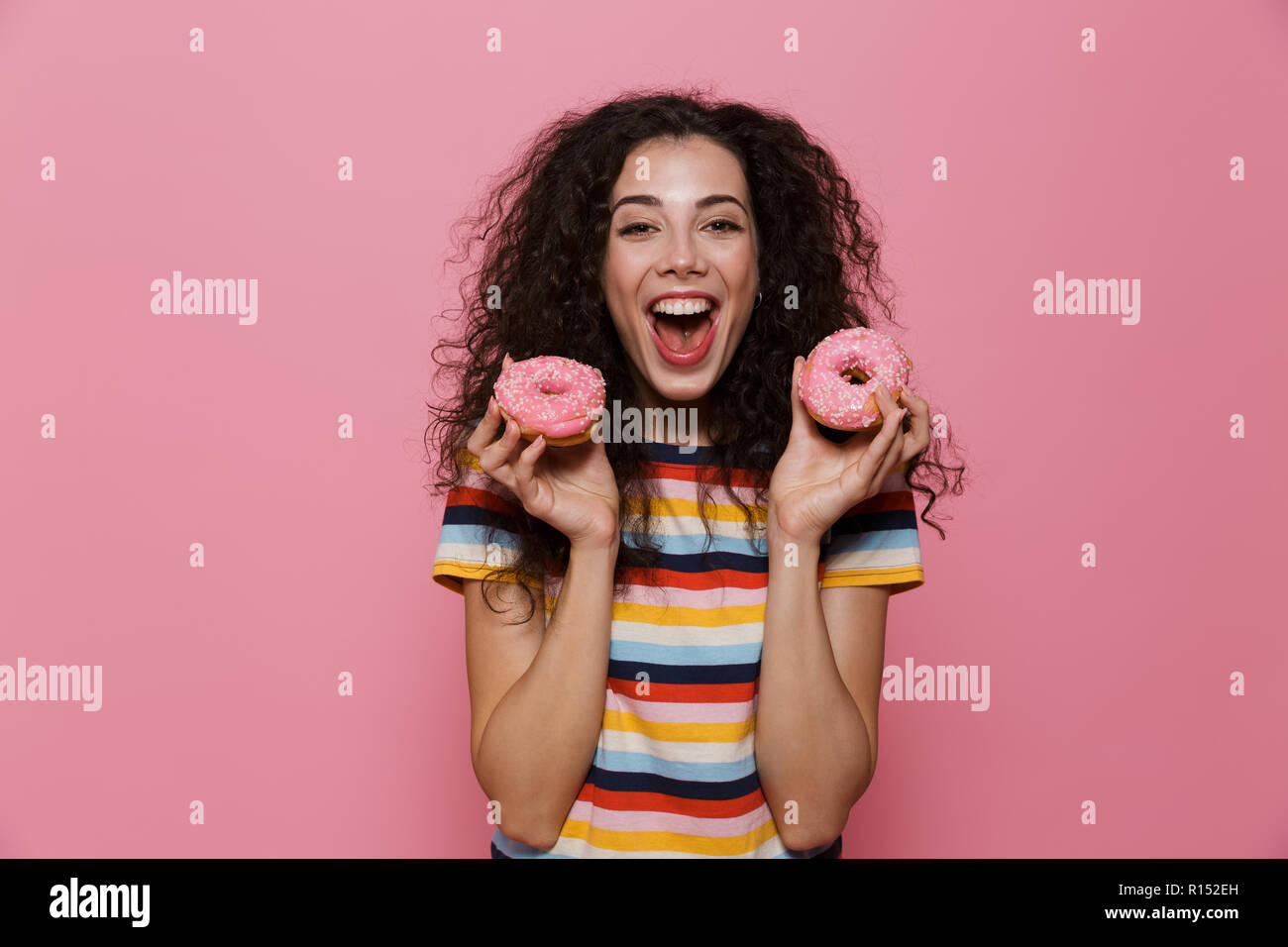 Photo de femme joyeuse 20s avec les cheveux bouclés s'amusant et holding donuts plus isolé sur fond rose Banque D'Images