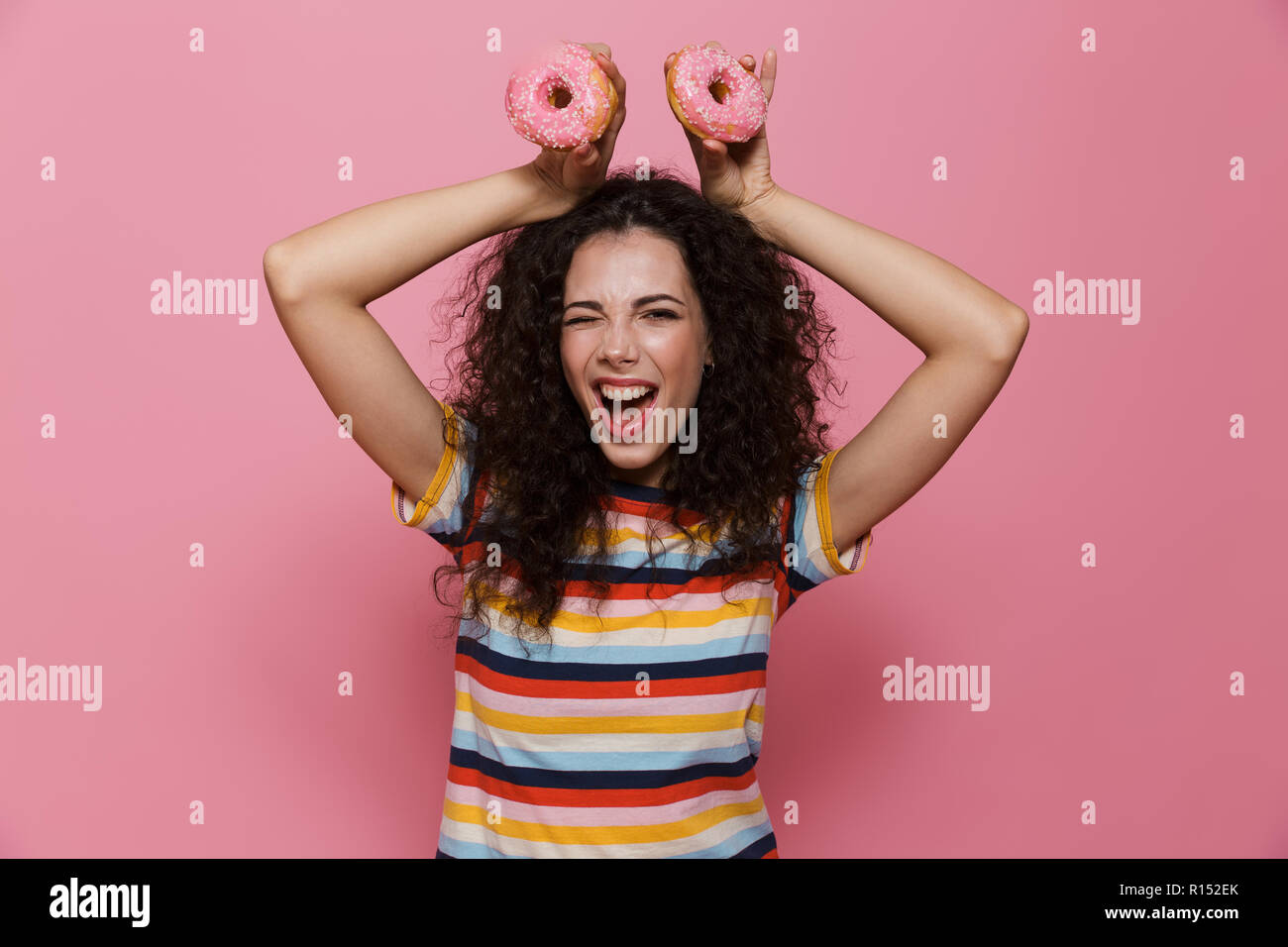 Photo de belle femme 20s avec les cheveux bouclés s'amusant et holding donuts plus isolé sur fond rose Banque D'Images
