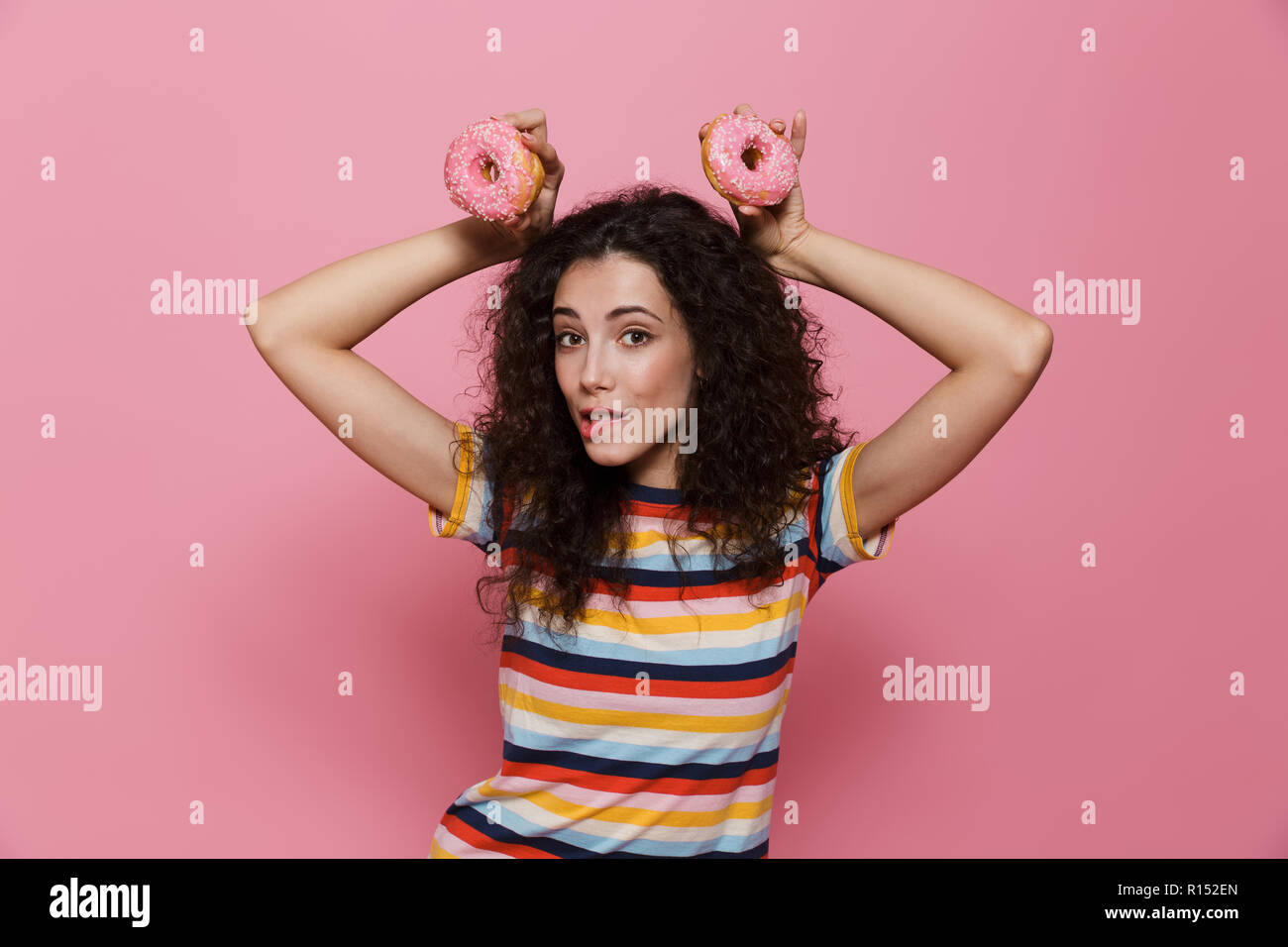 Photo de cute woman 20s avec les cheveux bouclés s'amusant et holding donuts plus isolé sur fond rose Banque D'Images