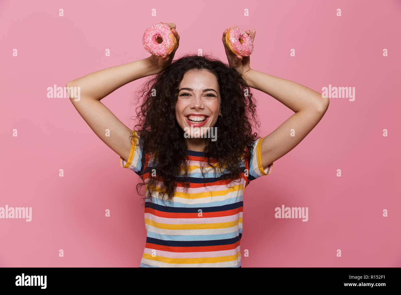 Photo de femme espiègle 20s avec les cheveux bouclés s'amusant et holding donuts plus isolé sur fond rose Banque D'Images