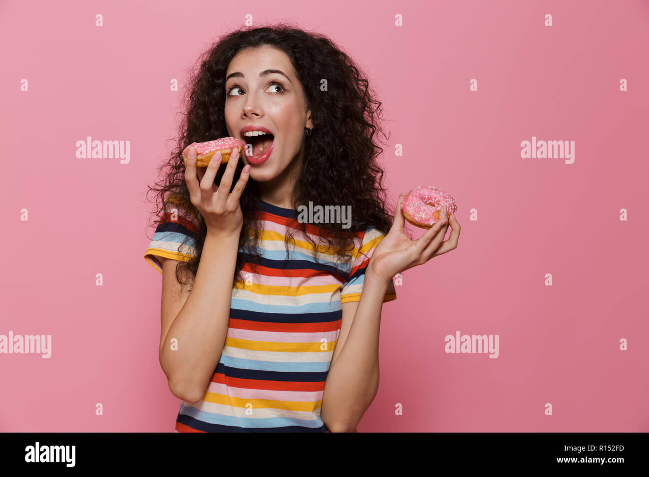 Photo de femme élégante 20s avec les cheveux bouclés s'amusant et holding donuts plus isolé sur fond rose Banque D'Images