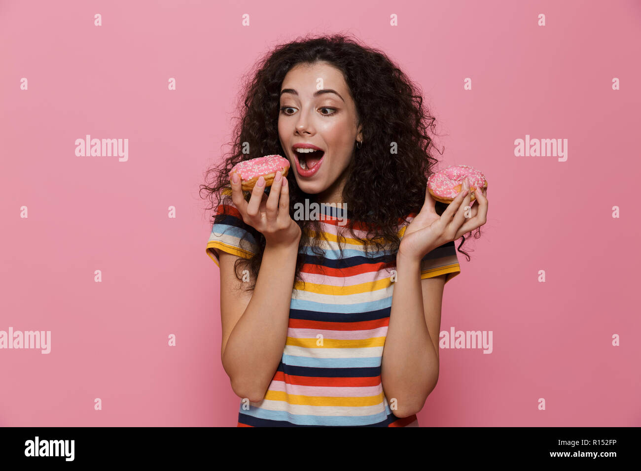 Photo de femme brune 20s avec les cheveux bouclés de jouer et de manger des beignes au fil isolé sur fond rose Banque D'Images