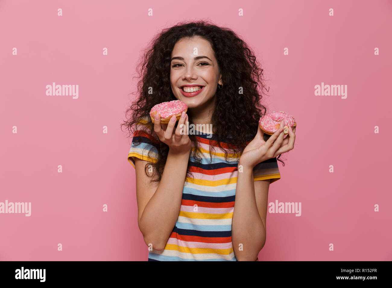 Photo de femme européenne 20s avec les cheveux bouclés de jouer et de manger des beignes au fil isolé sur fond rose Banque D'Images