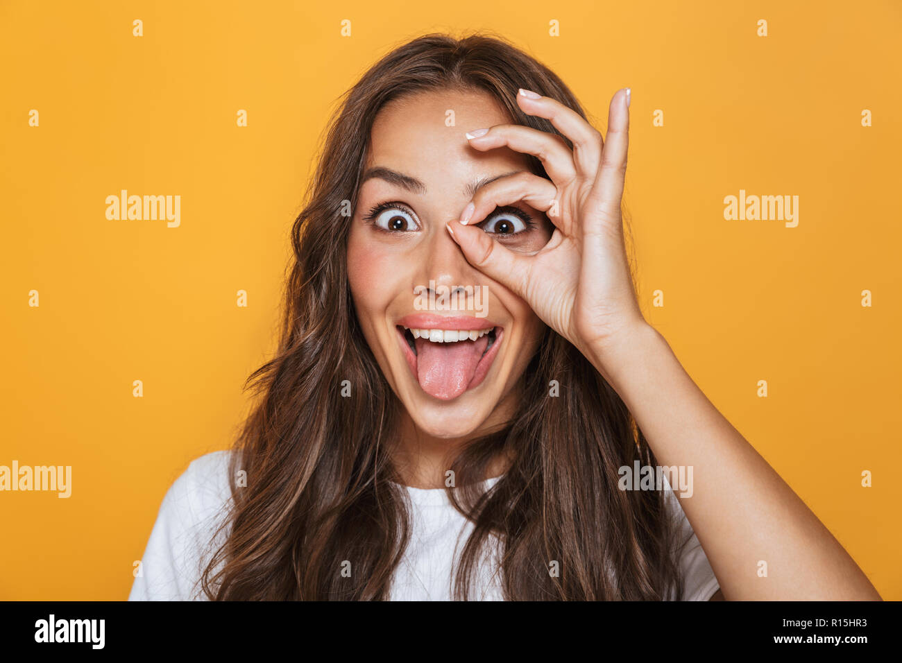 Portrait de femme brune 20s avec de longs cheveux de sourire et à la recherche à travers le trou fait avec ses doigts plus isolé sur fond jaune Banque D'Images
